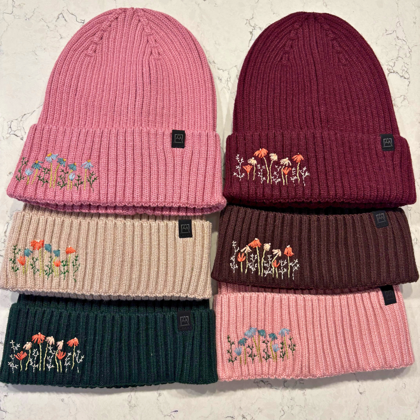 6 different colored beanies with dainty embroidered floral design in bottom left corner.