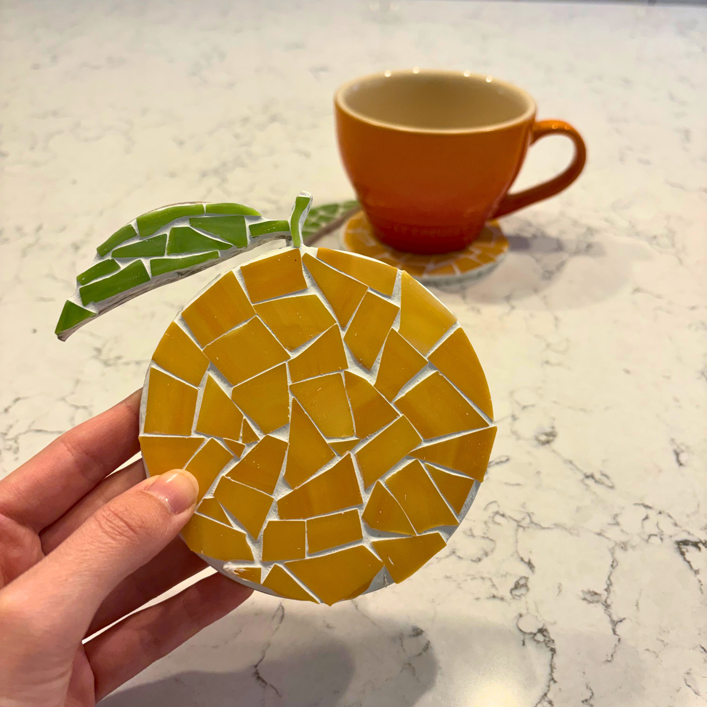 Hand holding an orange fruit mosaic coaster with a cup in the background on a marble surface