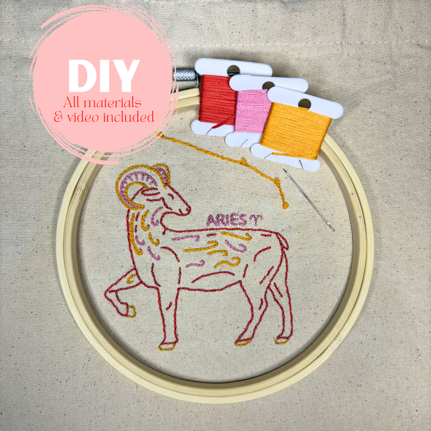 Embroidery hoop with Aries zodiac sign design and colored thread spools on a beige fabric background.