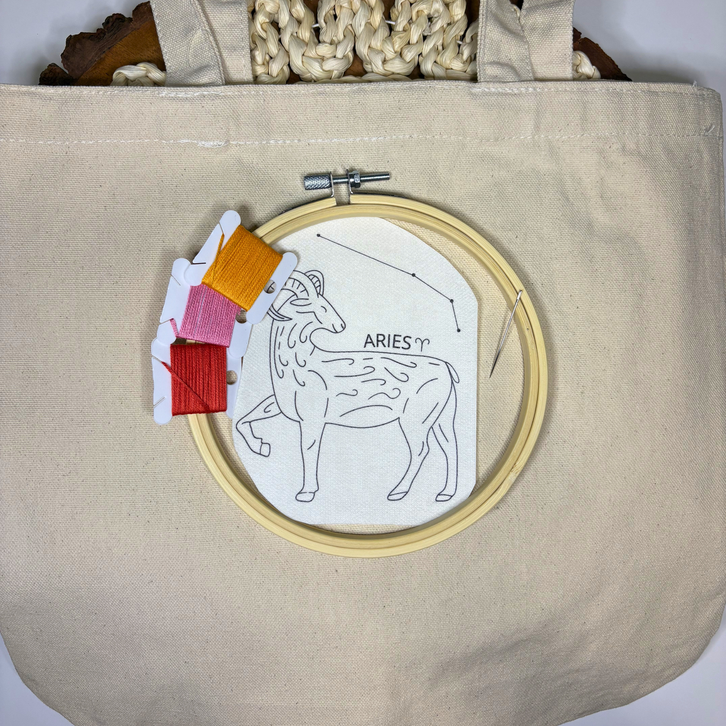 Aries zodiac design sticker with colorful embroidery thread, needle and hoop on top of a beige canvas tote bag.