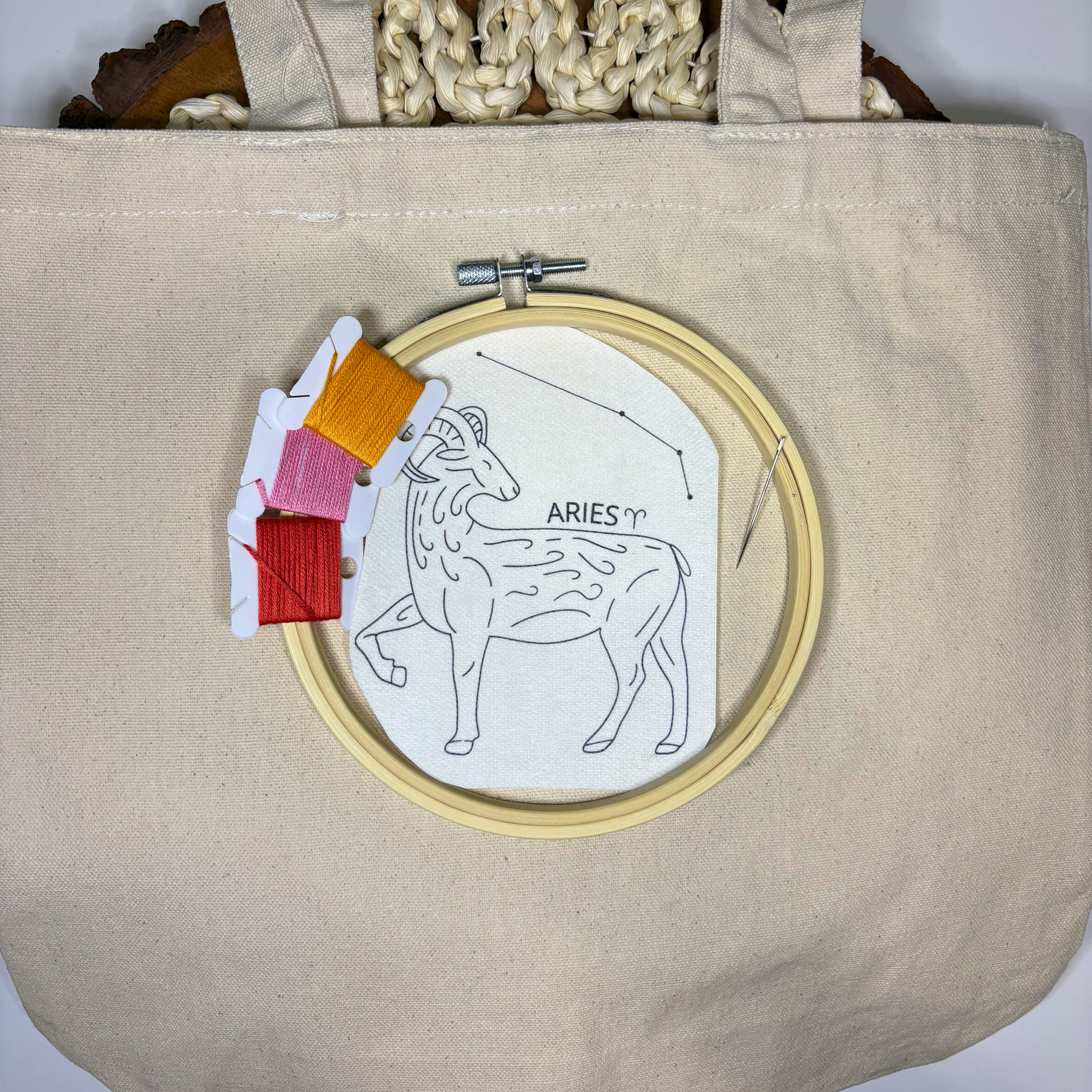 Aries zodiac design sticker with colorful embroidery thread, needle and hoop on top of a beige canvas tote bag.