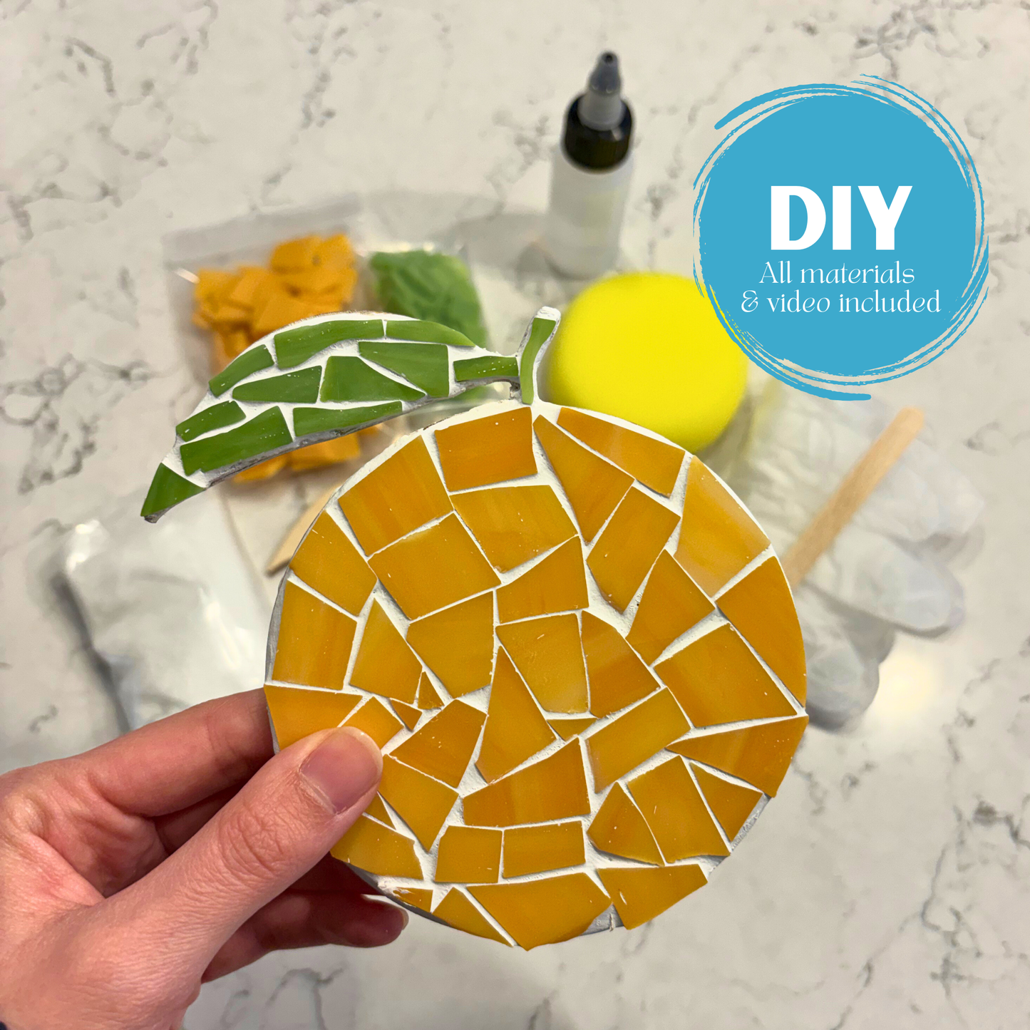 Hand holding a mosaic orange coaster with DIY text and materials on a marble surface.