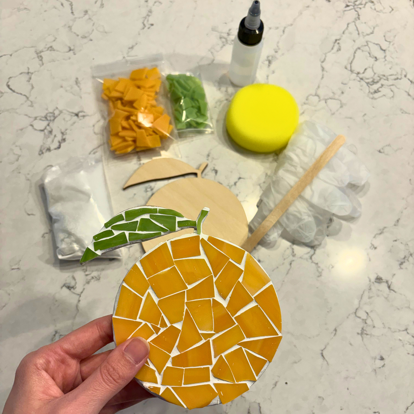 Hand holding a mosaic orange coaster with supplies in background on a marble countertop