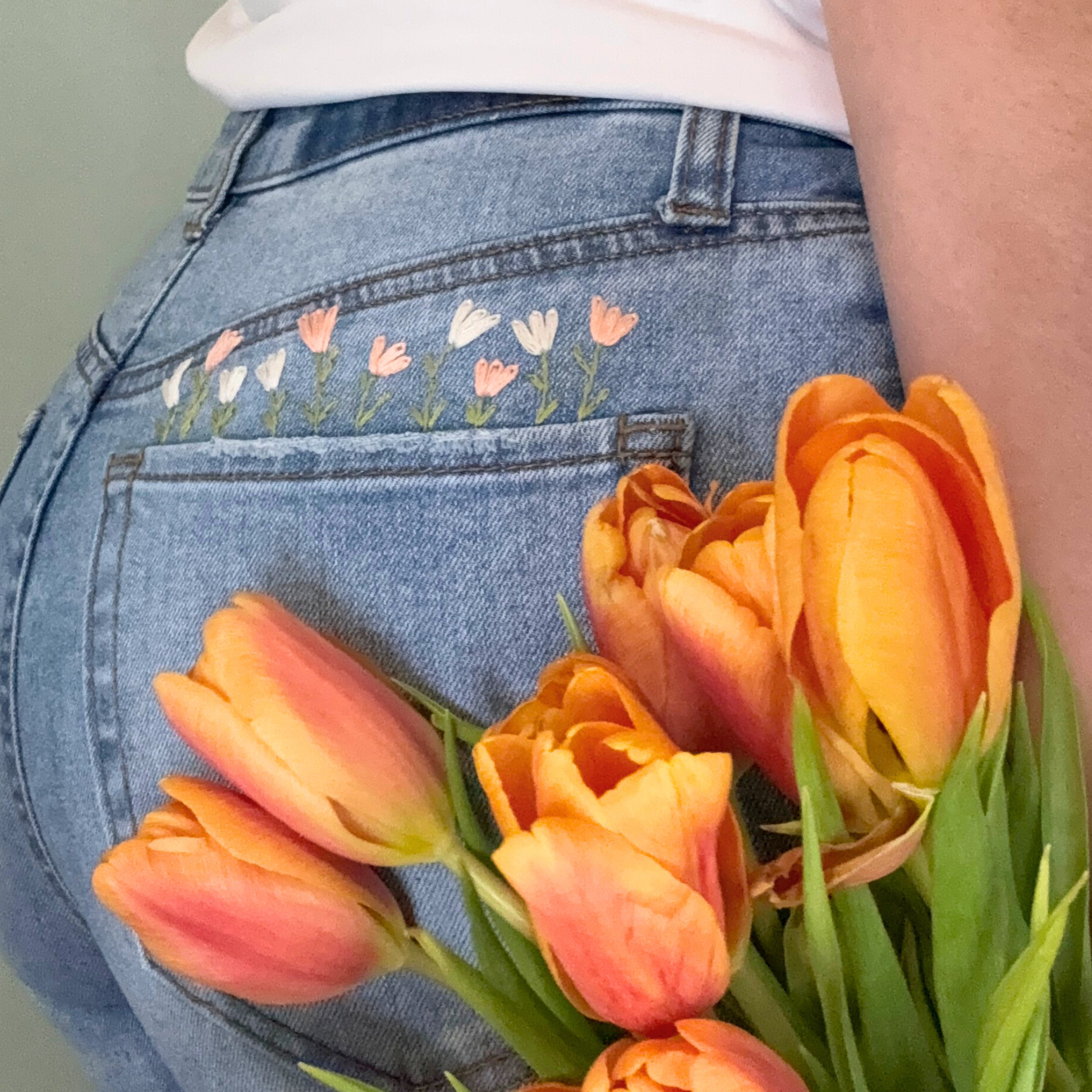 Person holding a bouquet of orange tulips with embroidered flowers on blue jeans.