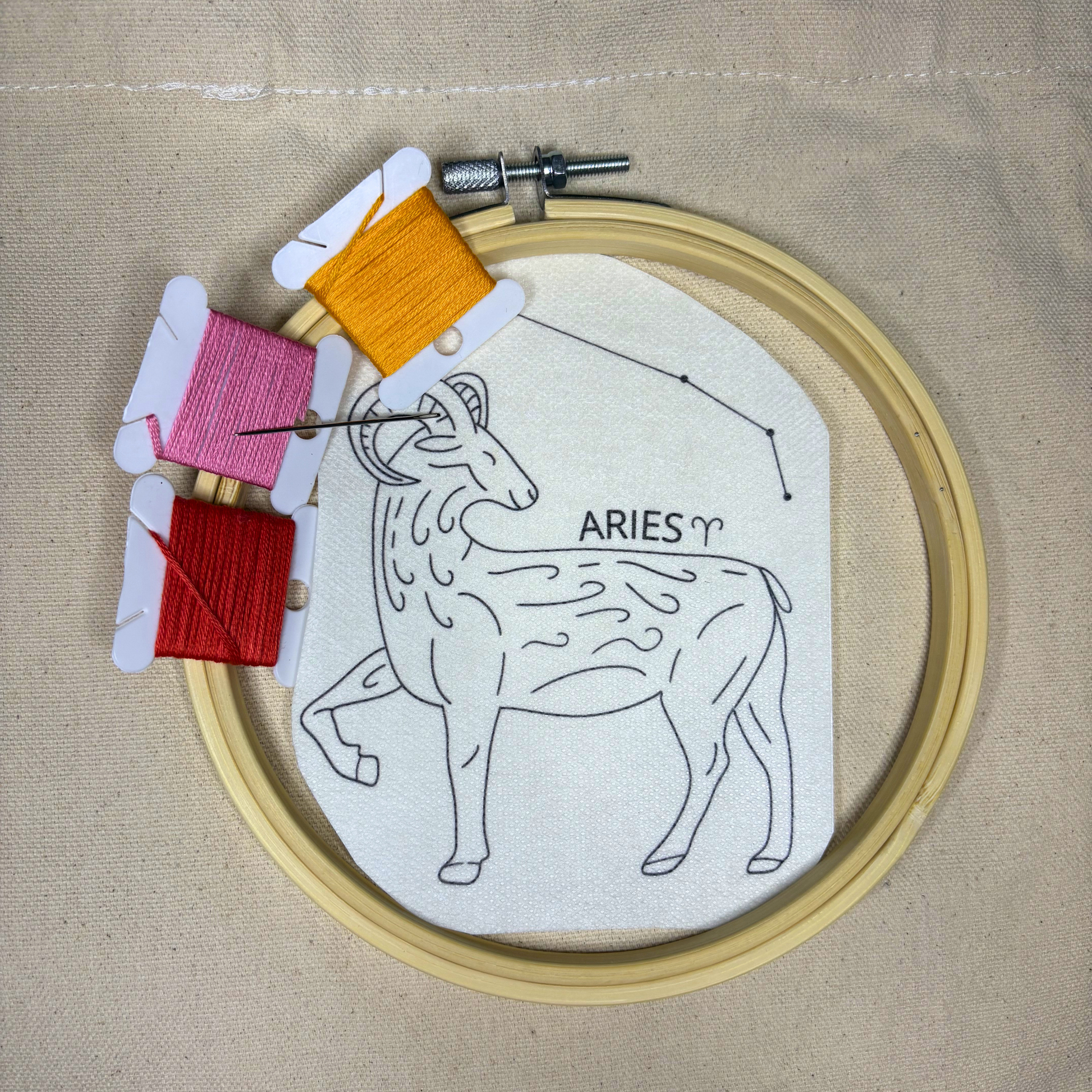 Aries zodiac design sticker with colorful embroidery thread with a hoop on top of a beige canvas tote bag.