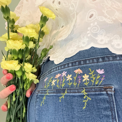Person holding yellow flowers with embroidered floral design on blue jeans and white lace top.