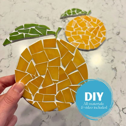 Hand holding a mosaic orange with another orange coaster in the background on a marble surface.