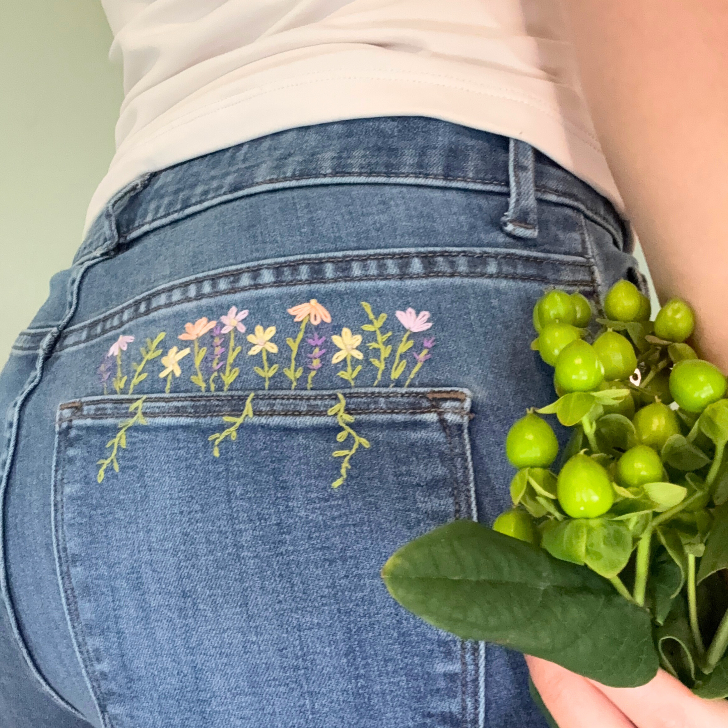 Person wearing jeans with floral embroidery holding green leaves.