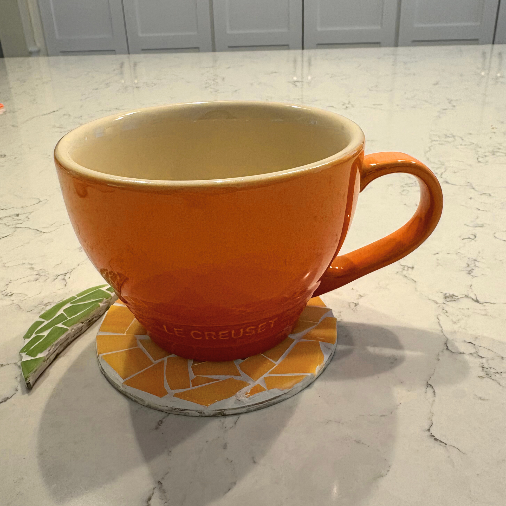 Orange Le Creuset mug on a mosaic coaster on a marble countertop