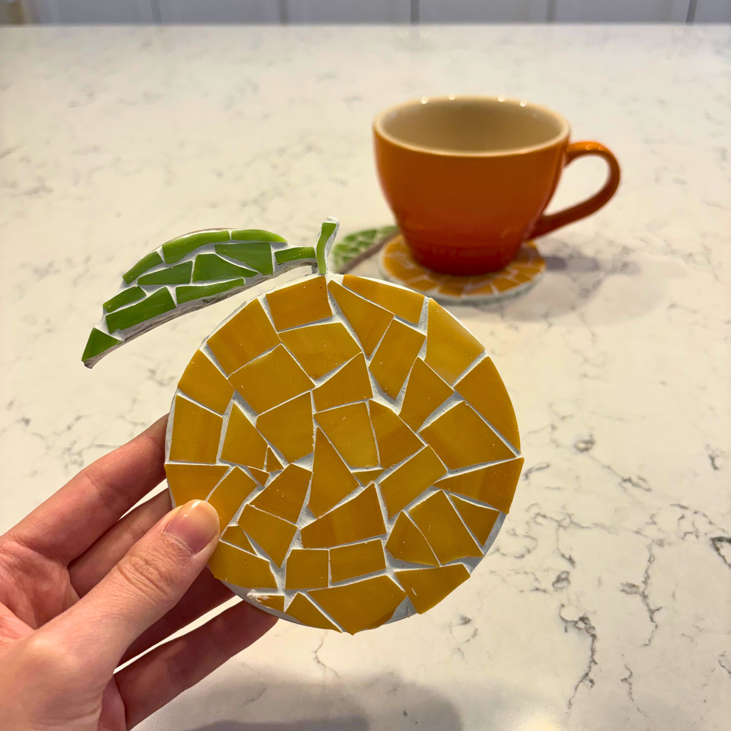 Hand holding a mosaic orange coaster with an orange mug in the background.