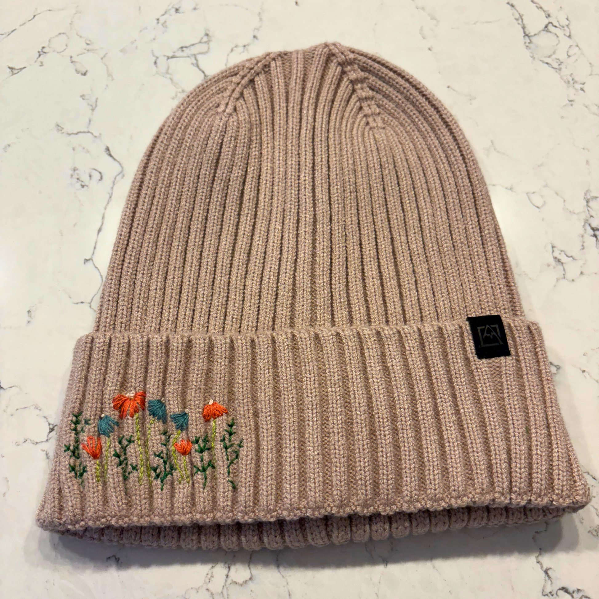 Light tan beanie with dainty embroidered floral design in bottom left corner.