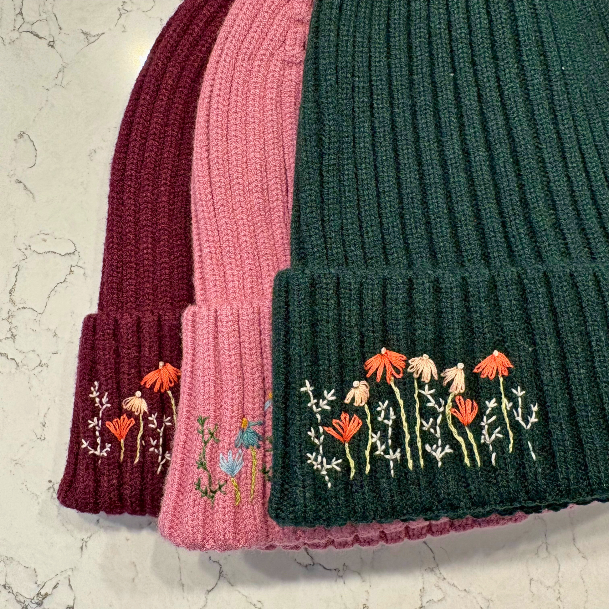 Three knitted hats in different colors with floral embroidery on a marble background